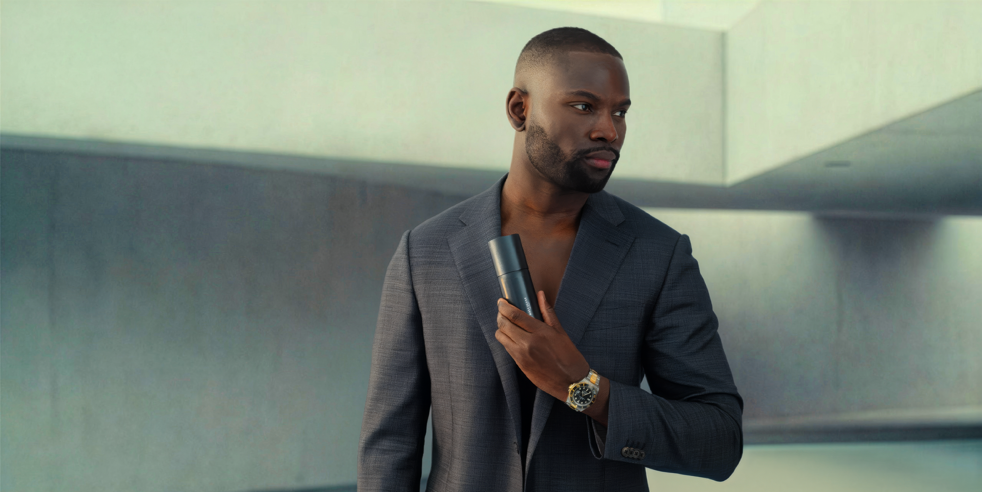 A man in a dark gray suit jacket holding a slim Masculuxe product, standing in a modern concrete hallway. He is wearing a gold-toned wristwatch.
